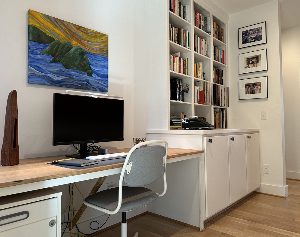 A painting of a green island in a blue ocean is above a desk and computer. A painting of a green island in a blue ocean is above a desk and computer.