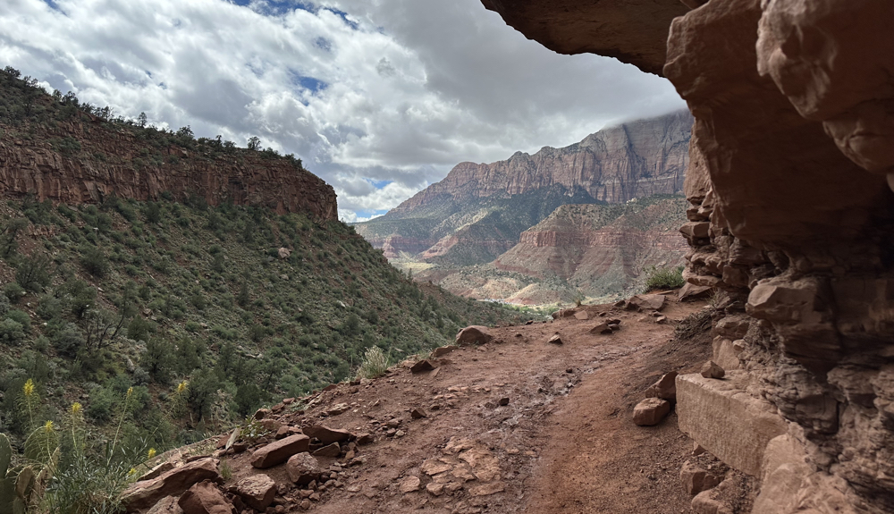 Trail bending around rocky cliff with valley and canyon walls in distance.